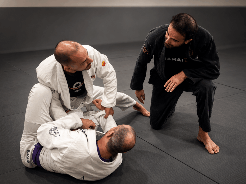 Jaime, de kimono, dando um seminário de Jiu-Jitsu e demonstrando uma técnica para dois alunos no tatame
